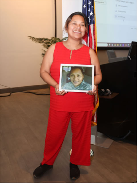 Woman in red holding a framed photo of a woman veteran in uniform beside the American flag